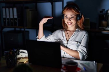Young caucasian woman working at the office at night gesturing with hands showing big and large size sign, measure symbol. smiling looking at the camera. measuring concept. 