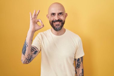 Hispanic man with tattoos standing over yellow background smiling positive doing ok sign with hand and fingers. successful expression. 