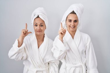 Middle age woman and daughter wearing white bathrobe and towel pointing finger up with successful idea. exited and happy. number one. 