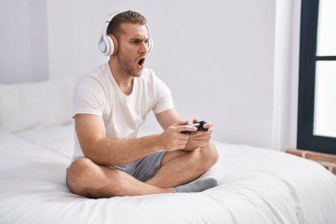 Young caucasian man sitting on the bed at home playing video games in shock face, looking skeptical and sarcastic, surprised with open mouth 