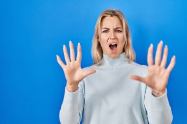 Young caucasian woman standing over blue background afraid and terrified with fear expression stop gesture with hands, shouting in shock. panic concept. 