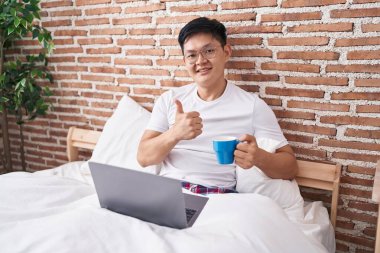 Young asian man drinking coffee sitting on the bed smiling happy and positive, thumb up doing excellent and approval sign 