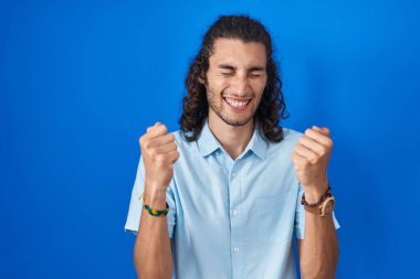 Mavi arka planda duran genç İspanyol adam başarı için heyecanlı kolları havada ve gözleri kapalı zaferi kutlarken gülümsüyor. Kazanan konsept. 