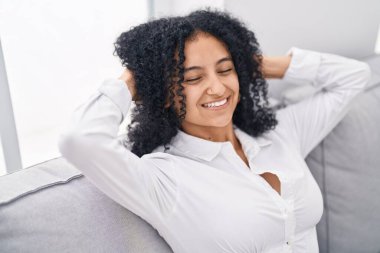 Young hispanic woman relaxed with hands on head sitting on sofa at home