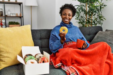 Afro-Amerikalı kadın kahvaltısını evde, kanepede yapıyor.