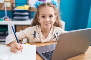 Young little girl sitting on the table doing homework with laptop looking positive and happy standing and smiling with a confident smile showing teeth 
