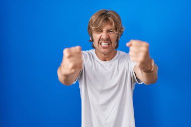 Middle age man standing over blue background angry and mad raising fists frustrated and furious while shouting with anger. rage and aggressive concept. 