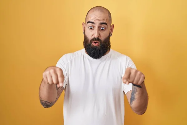 Young hispanic man with beard and tattoos standing over yellow background pointing down with fingers showing advertisement, surprised face and open mouth