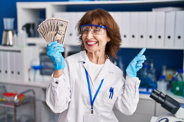 Middle age woman working at scientist laboratory holding money smiling happy pointing with hand and finger to the side