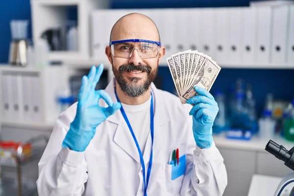 Young Hispanic Man Working Scientist Laboratory Holding Money Doing Sign — Stok fotoğraf