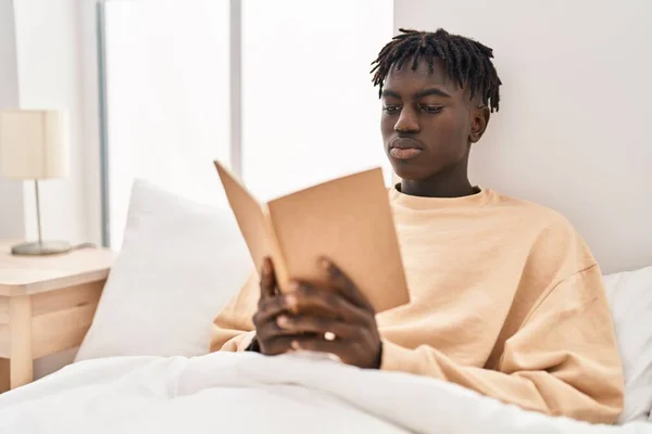 African american man reading book sitting on bed at bedroom