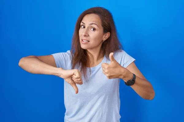 Brunette woman standing over blue background doing thumbs up and down, disagreement and agreement expression. crazy conflict