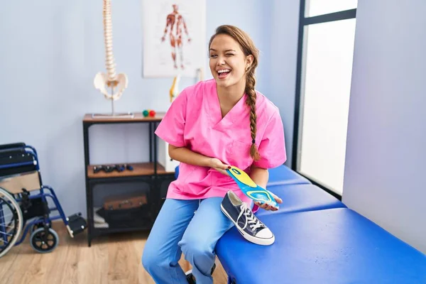 Young hispanic woman holding shoe insole at physiotherapy clinic winking looking at the camera with sexy expression, cheerful and happy face.