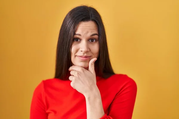 Young hispanic woman standing over yellow background looking confident at the camera smiling with crossed arms and hand raised on chin. thinking positive.