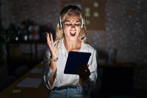 Young blonde woman working at the office at night crazy and mad shouting and yelling with aggressive expression and arms raised. frustration concept.