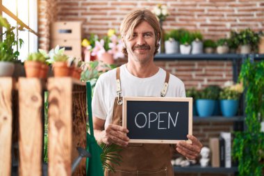 Caucasian man with mustache working at florist holding open sign relaxed with serious expression on face. simple and natural looking at the camera. 