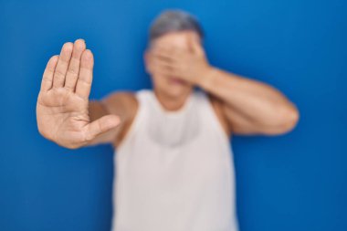 Orta yaşlı, beyaz bir kadın mavi arka planda dikiliyor, elleriyle gözlerini kapatıyor ve üzgün ve korku dolu bir ifadeyle dur hareketi yapıyor. utangaç ve olumsuz kavram. 