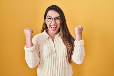 Young hispanic woman standing over yellow background celebrating surprised and amazed for success with arms raised and open eyes. winner concept. 