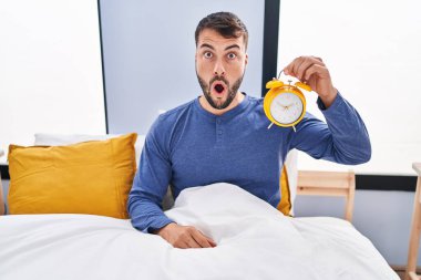 Handsome hispanic man in the bed holding alarm clock scared and amazed with open mouth for surprise, disbelief face 