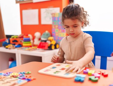 Adorable hispanic girl playing with maths puzzle game sitting on table at kindergarten