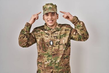 Genç Arap adam kamuflaj üniforması giyiyor. İki eliyle başını gösteren gülümsüyor. Harika bir fikir ya da düşünce. 