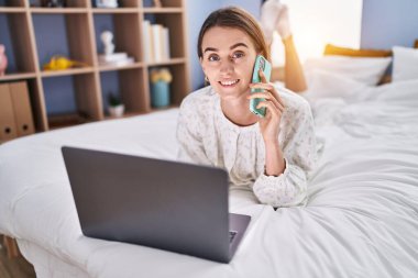 Young caucasian woman using laptop talking on smartphone at bedroom