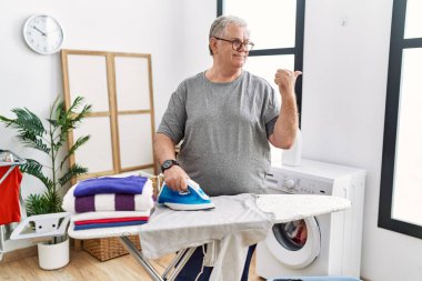 Senior caucasian man ironing clothes at home pointing thumb up to the side smiling happy with open mouth 