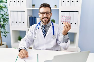 Handsome hispanic man wearing doctor uniform holding prescription pills looking positive and happy standing and smiling with a confident smile showing teeth 