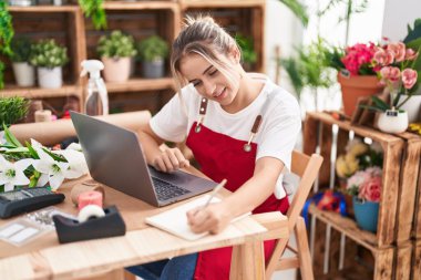 Young blonde woman florist using laptop writing on notebook at flower shop