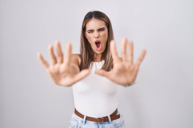 Hispanic young woman standing over white background doing stop gesture with hands palms, angry and frustration expression 