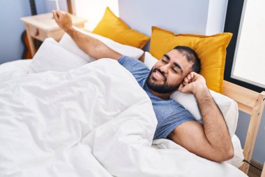 Young hispanic man waking up stretching arms at bedroom