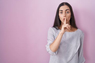 Genç esmer kadın pembe arka planda dikiliyor ve parmakları dudaklarında sessiz olmak istiyor. sessizlik ve gizli konsept. 