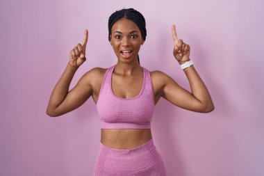 African american woman with braids wearing sportswear over pink background smiling amazed and surprised and pointing up with fingers and raised arms. 