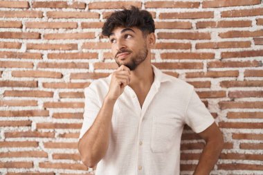 Arab man with beard standing over bricks wall background with hand on chin thinking about question, pensive expression. smiling with thoughtful face. doubt concept. 