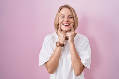 Pembe arka planda duran genç beyaz kadın ağzı açık gülümsüyor, parmakları işaret ediyor ve neşeli bir gülümseme sergiliyor. 