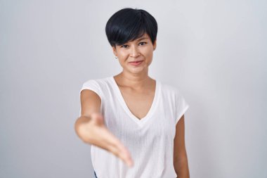 Young asian woman with short hair standing over isolated background smiling friendly offering handshake as greeting and welcoming. successful business. 