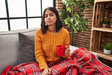 Young hispanic woman sitting on the sofa drinking a coffee at home looking sleepy and tired, exhausted for fatigue and hangover, lazy eyes in the morning. 