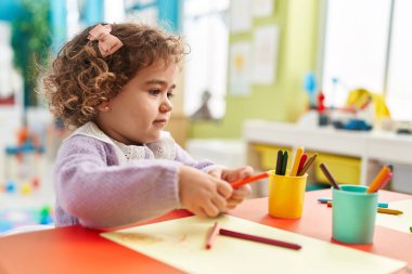 Adorable hispanic girl preschool student sitting on table drawing on paper at kindergarten