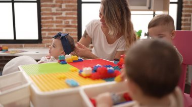Öğretmen ve anaokulu öğrencileri anaokulundaki masada oturan inşaat bloklarıyla oynuyorlar.