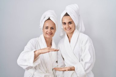 Middle age woman and daughter wearing white bathrobe and towel gesturing with hands showing big and large size sign, measure symbol. smiling looking at the camera. measuring concept. 