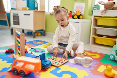 Adorable blonde girl playing with maths puzzle game sitting on floor at kindergarten