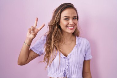 Pembe arka planda duran genç İspanyol kadın iki numarayı gösterip işaret ederken kendinden emin ve mutlu bir şekilde gülümsüyor.. 