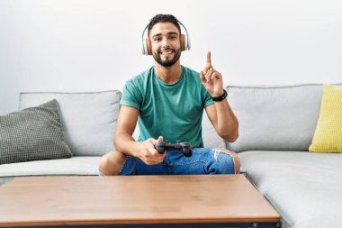 Handsome hispanic man wearing headphones playing video game holding controller surprised with an idea or question pointing finger with happy face, number one 