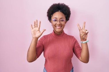 Beautiful african woman with curly hair standing over pink background showing and pointing up with fingers number seven while smiling confident and happy. 