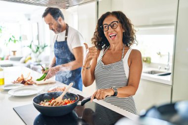 Middle age couple cooking mediterranean food at home pointing thumb up to the side smiling happy with open mouth 