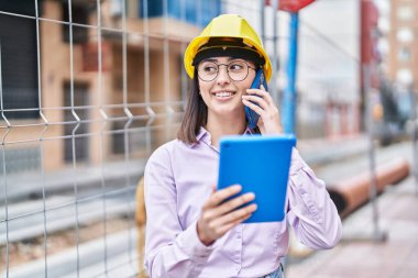 Young hispanic woman architect talking on the smartphone using touchpad at street
