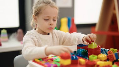 Adorable blonde girl playing with construction blocks sitting on table at kindergarten