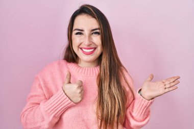 Pembe arka planda duran genç İspanyol kadın avuç içini gösteriyor ve baş parmaklarıyla iyi bir hareket yapıyor, mutlu ve neşeli gülümsüyor. 