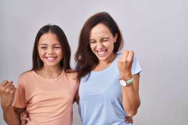 Young mother and daughter standing over white background very happy and excited doing winner gesture with arms raised, smiling and screaming for success. celebration concept. 
