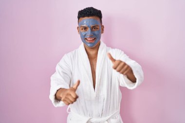 Young hispanic man wearing beauty face mask and bath robe approving doing positive gesture with hand, thumbs up smiling and happy for success. winner gesture. 
